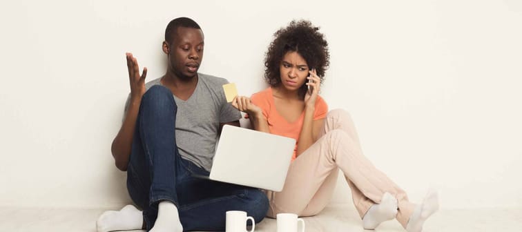 Sad black couple shopping online on laptop, making order on mobile, paying with credit card while sitting at home floor in empty living room, copy space, isolated