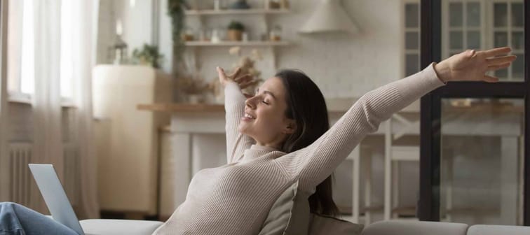 Happy young woman relaxing alone on cozy sofa put wireless computer on laps, raising her arms