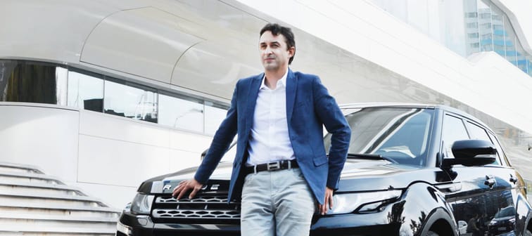 man in suit jacket leans against front of parked range rover in front of concrete building