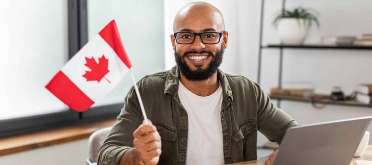 Happy latin man sitting with flag of Canada and using laptop computer, sitting at desk at home. Modern online foreign education, emigration and citizenship, working abroad concept
