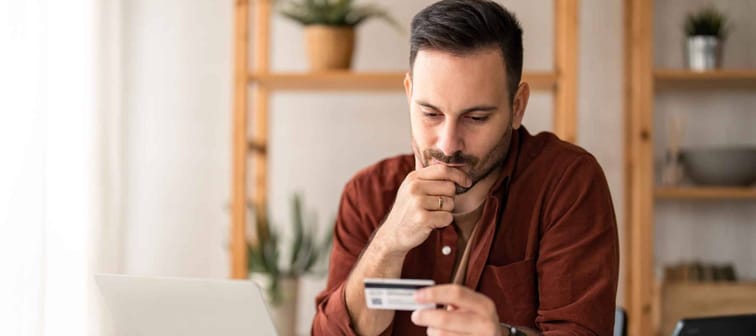 Serious business man sitting at table in home office, holding and looking at credit card, worried about debt and credit card deficit, considering a financial loan from bank.