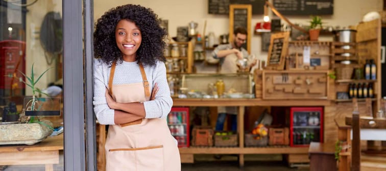 Portrait of a smiling young African American barista leaning with her arms crossed on the door of a trendy cafe