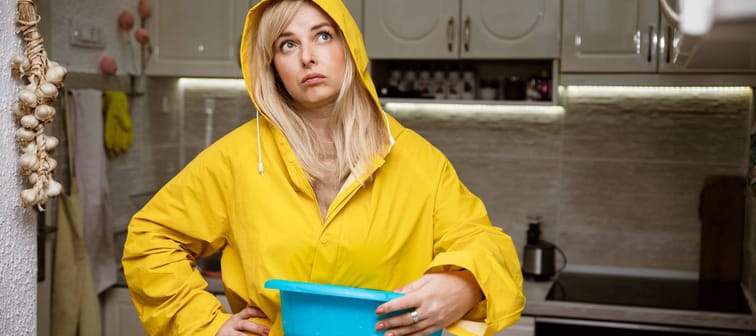 Woman in yellow raincoat holding bucket to catch rain inside home