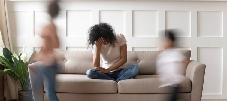 Overwhelmed mother sits on the couch as kids run around the room