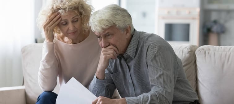 Worried senior wife and husband sit on sofa hold papers