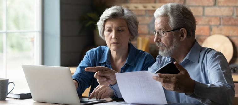 Focused senior husband and wife sit at table at home look at laptop screen.