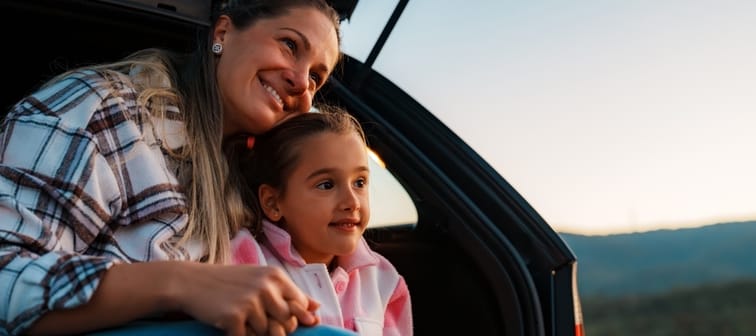 Mother and daughter are sitting in the open trunk of their car, holding hands and admiring a beautiful sunset over a picturesque landscape during a relaxing road trip.