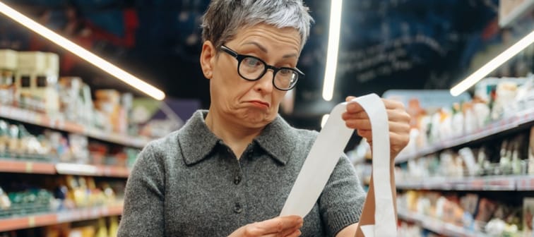 Older woman with short gray hair and glasses looks at her shopping receipt while holding an orange basket in a grocery store aisle filled with products.