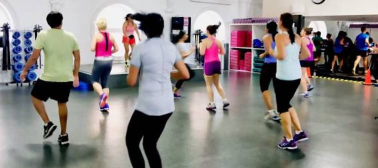 Group of people participating a workout class, backs to camera, facing instructor on a stage.