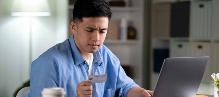 Serious young Asian man, frowning and worried while using and looking at credit card, doing online shopping, making online payment transaction using laptop  at home.