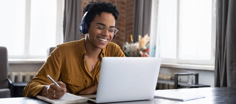 Cheerful Black college student in earphones attending virtual class, writing notes, watching webinar on Internet, studying online.