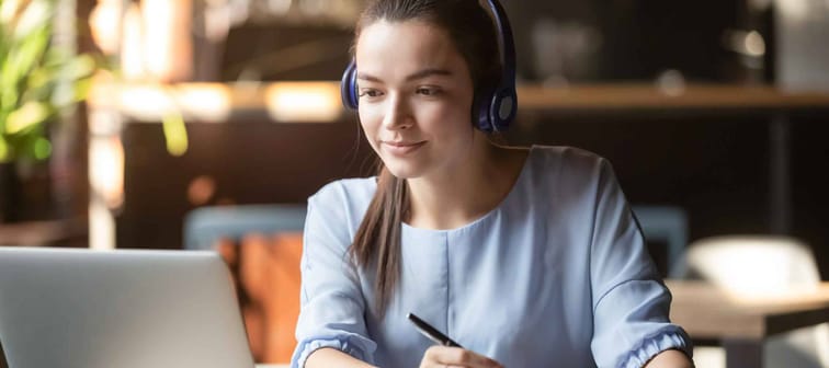 Focused woman wearing headphones using laptop in cafe, writing notes,  e-learning education concept