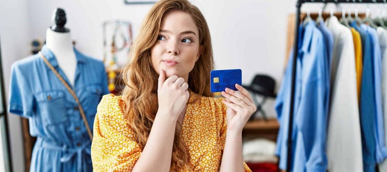 Young caucasian woman at retail shop holding credit card serious face thinking about question with hand on chin, thoughtful about confusing idea