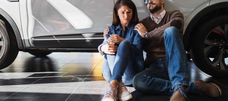 A sad young couple sits next to a car in a car dealership because they can't afford the car they want.