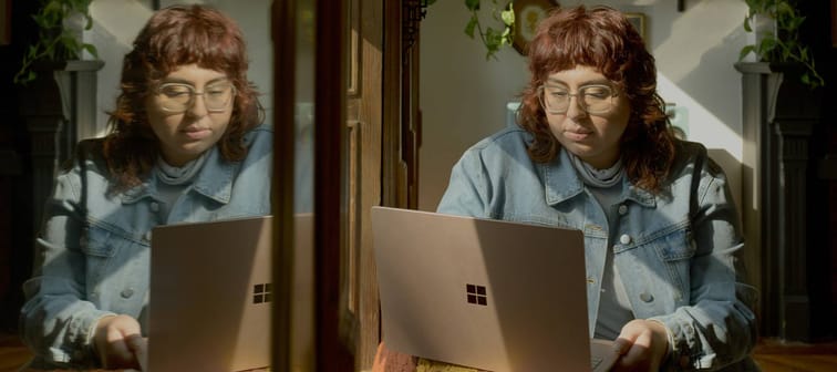 Person sitting at desk beside mirror showing their reflection as they work on their laptop
