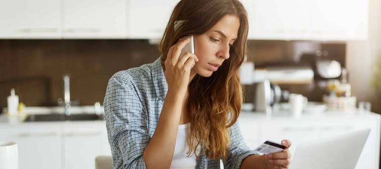 Caucasian woman calling bank using cell phone concerning information on credit card that she is holding. Serious female connecting to mobile banking service using electronic device