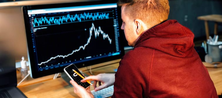 young man sits at desk tapping on cell phone with big monitor showing stock graph in backgroundd