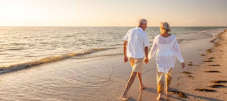 Happy senior man and woman old retired couple walking and holding hands on a beach at sunset, s3niorlife