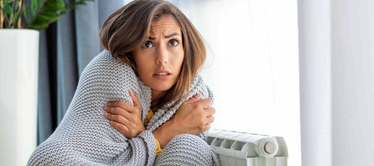 Unwell woman renter in blanket sit in cold living room hand on old radiator.suffer from lack of heat . Unhealthy young woman struggle from chill freeze at home. No heating concept.