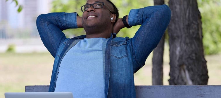 Happy black guy sits on a park bench waiting for his first date. A dreamy African-American man with laptops on his lap sat down to rest while waiting for a courier