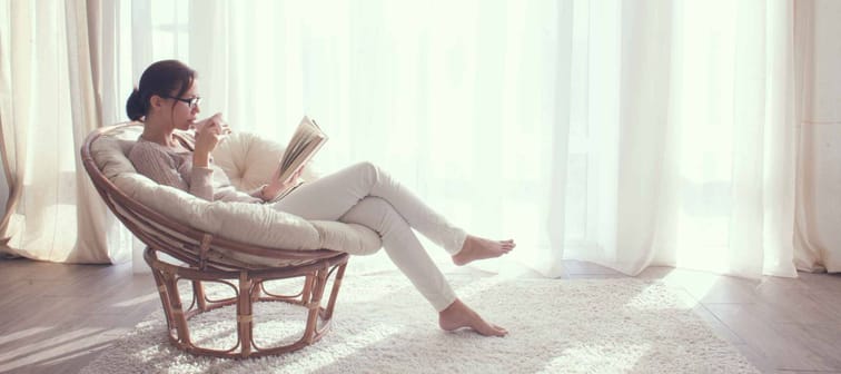 Young woman at home sitting on modern chair in front of window relaxing in her living room reading book and drinking coffee or tea