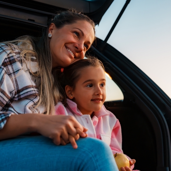 Mother and daughter are sitting in the open trunk of their car, holding hands and admiring a beautiful sunset over a picturesque landscape during a relaxing road trip.