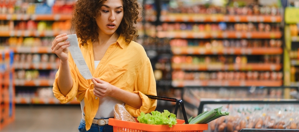 Woman holding a long shopping receipt, looking surprised and concerned about increasing food prices while shopping in a supermarket