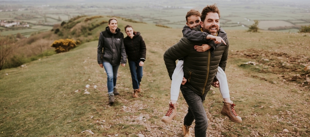 A joyful family hike features a man carrying a girl on his back, two women walk alongside.