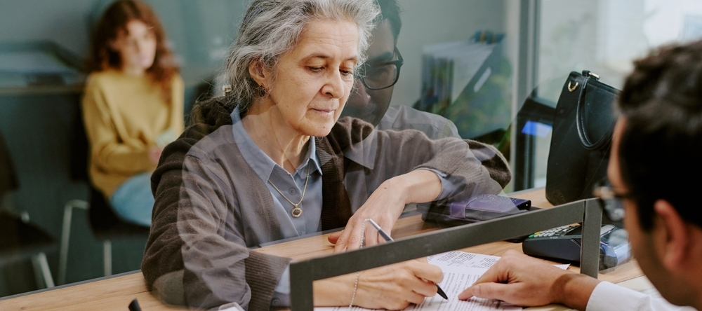Elderly woman signing official documents at banking counter with teller assisting