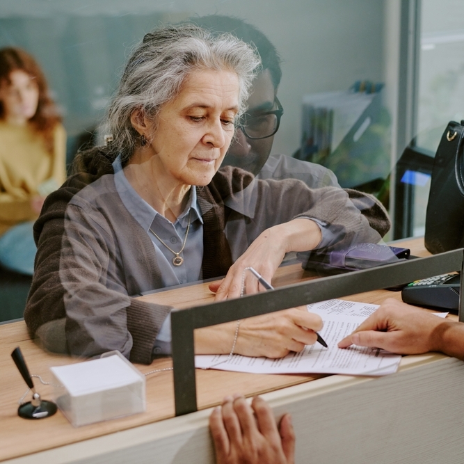 Elderly woman signing official documents at banking counter with teller assisting