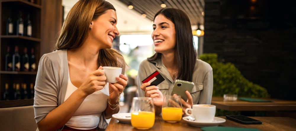Two female friends enjoy a coffee out while paying by credit card