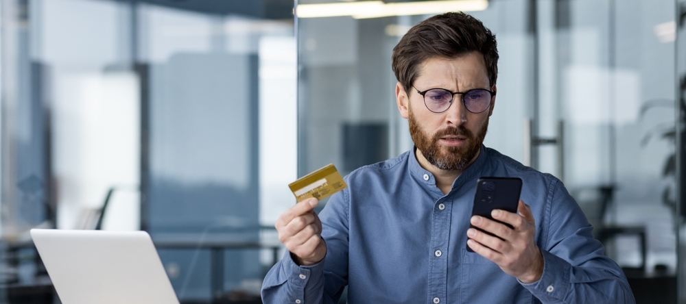 A man holding his credit card sitting at a desk with his laptop looking confused