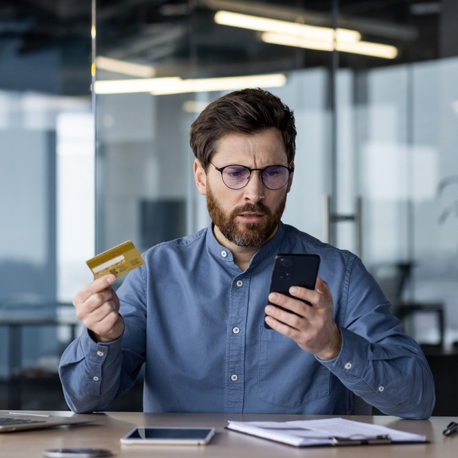 A man holding his credit card sitting at a desk with his laptop looking confused