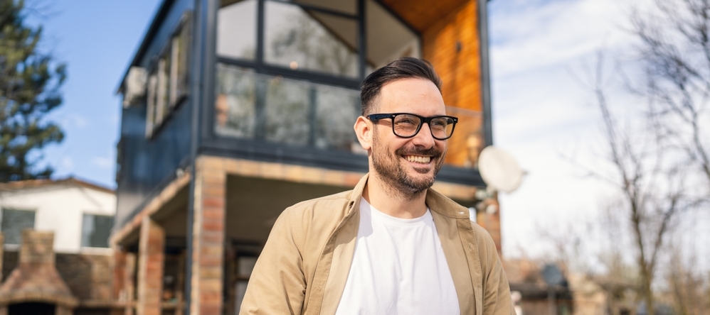 Man wearing glasses holding a coffee cup and standing in front of a house smiling.