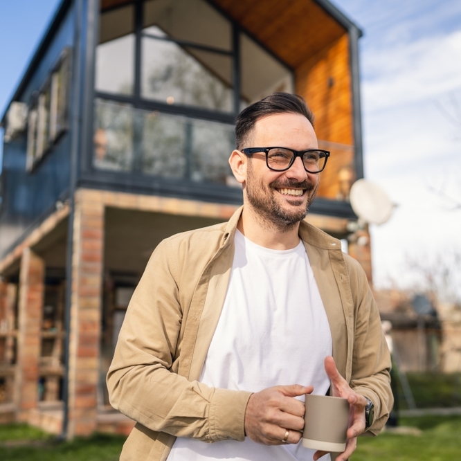 Man wearing glasses holding a coffee cup and standing in front of a house smiling.