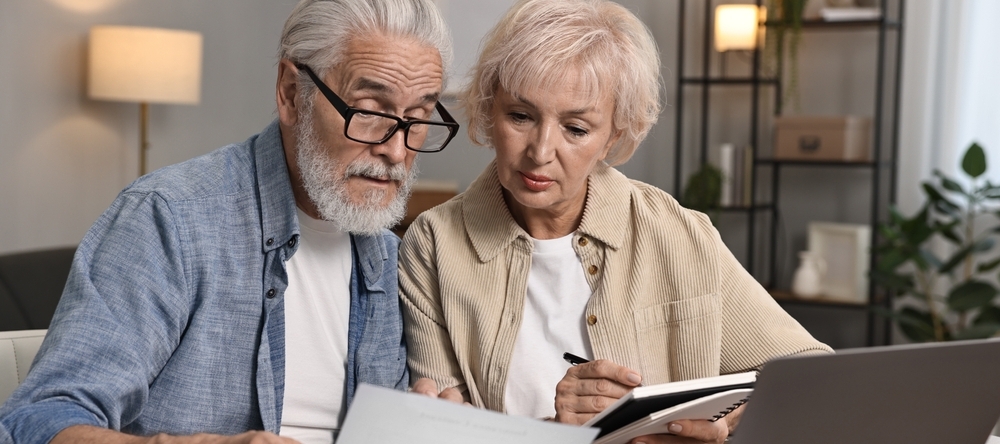 Retired male/female couple sitting at kitchen table looking over financial papers with a calculator and laptop nearby