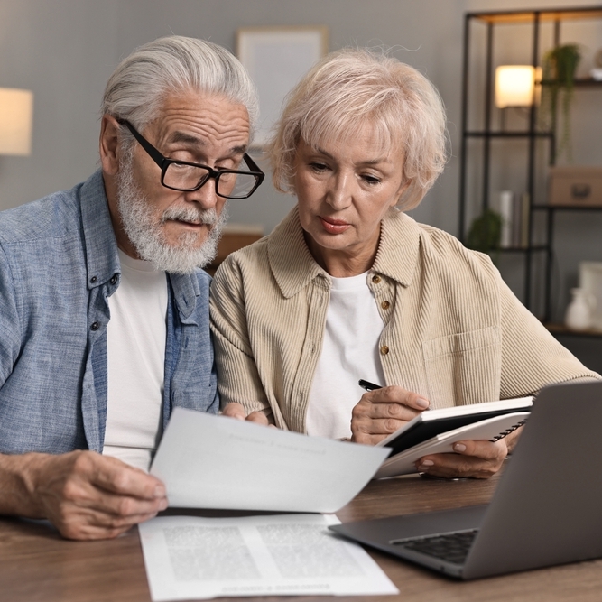 Retired male/female couple sitting at kitchen table looking over financial papers with a calculator and laptop nearby