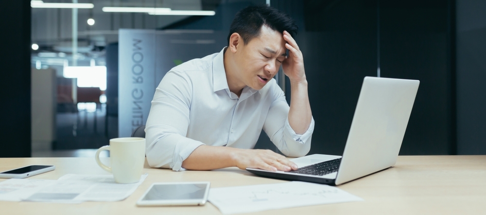 Professional Asian man sitting at desk looking stressed with a laptop in front of him
