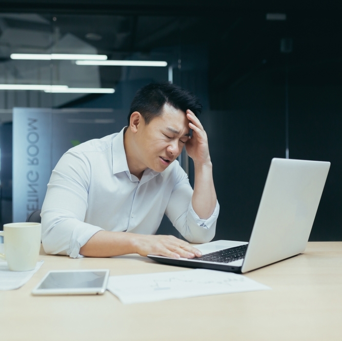 Professional Asian man sitting at desk looking stressed with a laptop in front of him