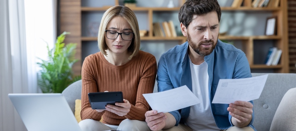 Stressed couple analyzing financial bills and expenses, using a calculator and laptop to manage debt and household budget, feeling worried about their money situation