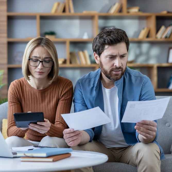Stressed couple analyzing financial bills and expenses, using a calculator and laptop to manage debt and household budget, feeling worried about their money situation