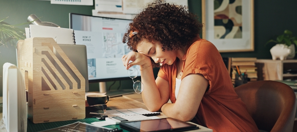 Woman sitting on desk, holding her glasses looking stressed