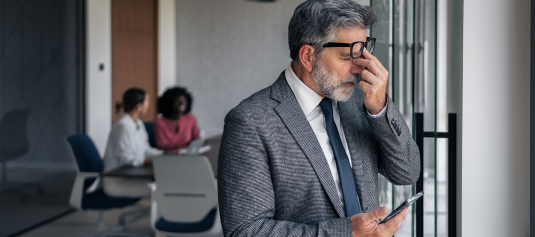 Mature businessman experiencing burnout and headache while holding phone in modern office, rubbing eyes due to strain with colleagues working in background, representing corporate pressure