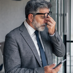 Mature businessman experiencing burnout and headache while holding phone in modern office, rubbing eyes due to strain with colleagues working in background, representing corporate pressure