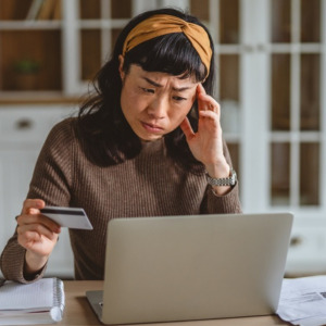 Woman feeling overwhelmed and stressed while trying to manage finances, looking intently at her laptop screen and holding a credit card to make payments at home
