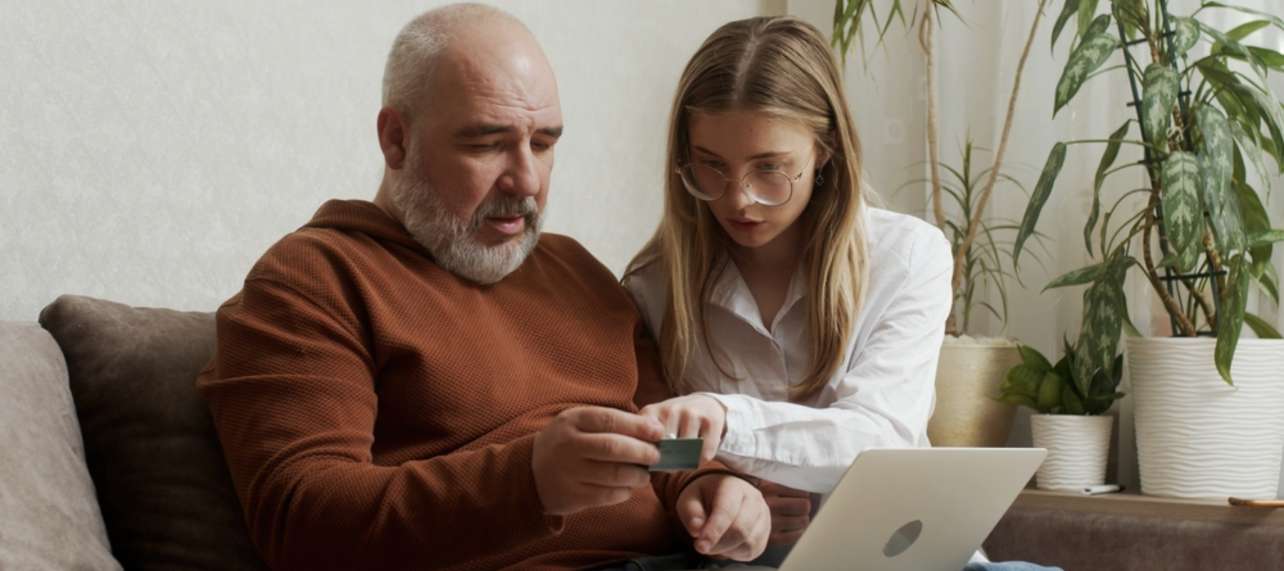 A pretty young woman shows her older mature father how to use a bank card and computer at home.