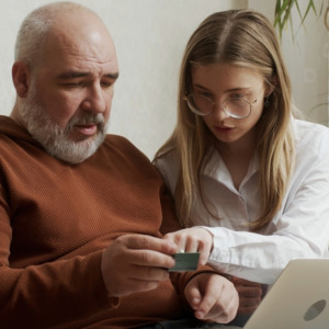 A pretty young woman shows her older mature father how to use a bank card and computer at home.