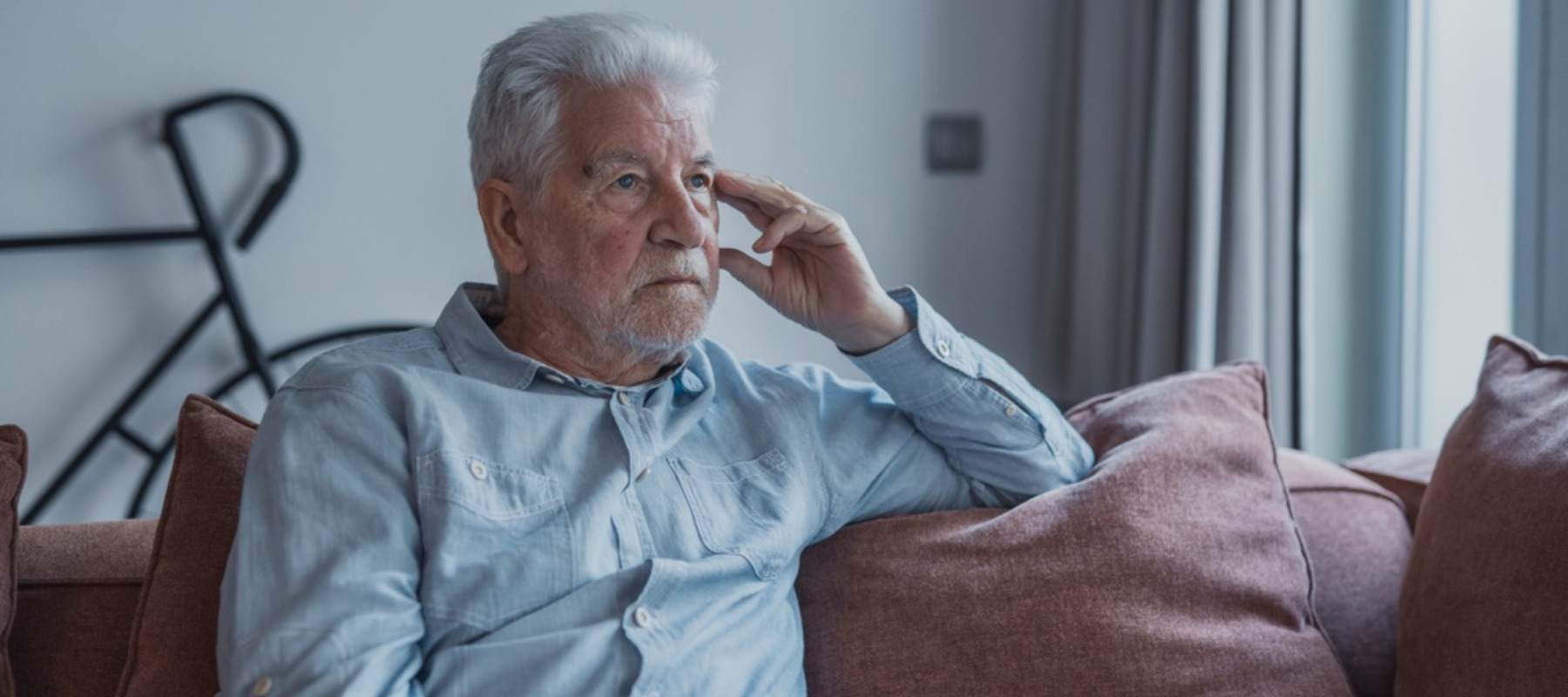 Elderly man sits alone on a sofa looking outside with a sad expression. He seems stressed and reflective while dealing with personal concerns at home.