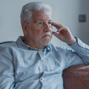 Elderly man sits alone on a sofa looking outside with a sad expression. He seems stressed and reflective while dealing with personal concerns at home.