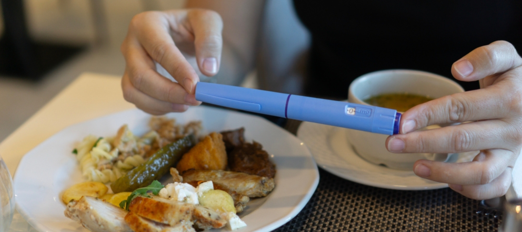 Close-up of hands holding an Ozempic injection pen while sitting at a table with a plate of food and a cup of soup